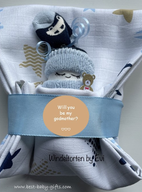 a blue diapered baby in a burp cloth with a sticker that says: will you be my godmother?